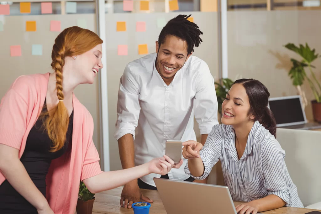 A diverse group of three young people laughing at something displayed on a phone in an office.