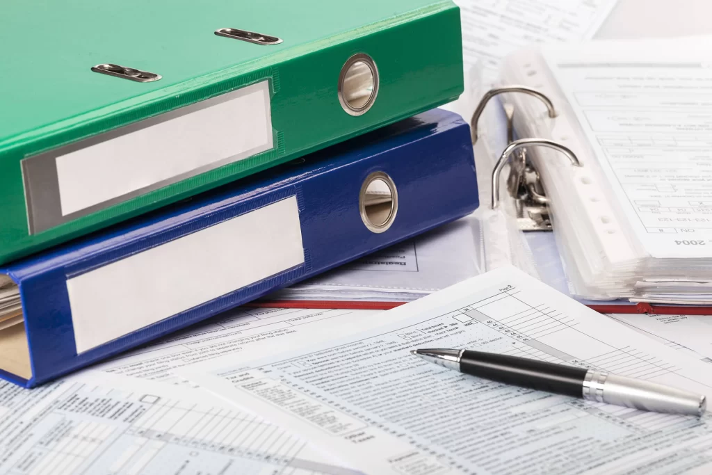 A desk full of binders and papers. A green binder on top of a blue binder on top of an open red binder with tax forms inside.