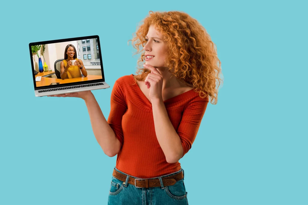 A red-haired woman holding an open laptop, which displays a Black woman behind a desk holding a coffee cup