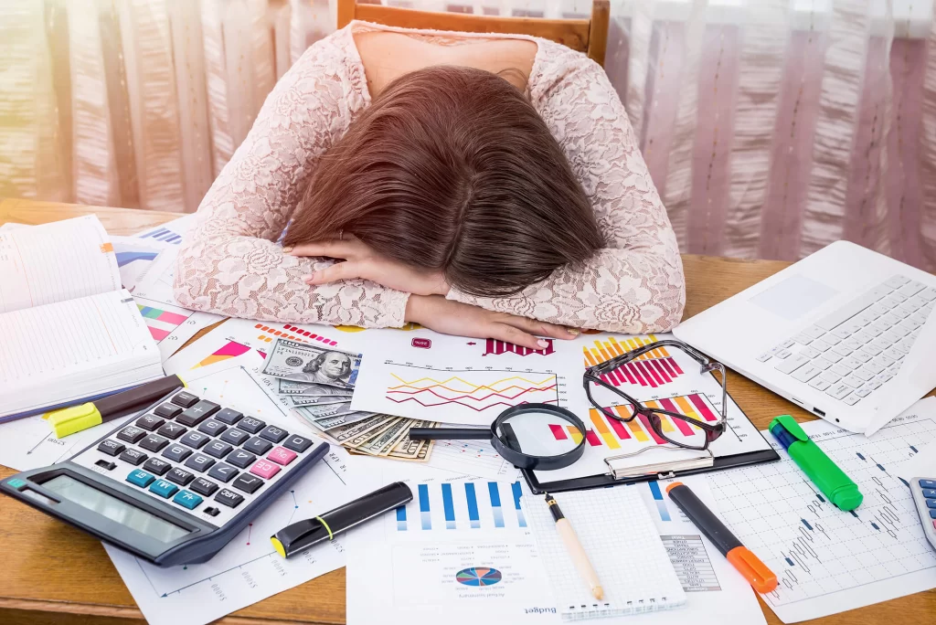 A woman rests her head on her arms at table covered in papers displaying different charts and graphics, as well as pens, highlighters, a calculator, a magnifying glass, glasses, and $100.