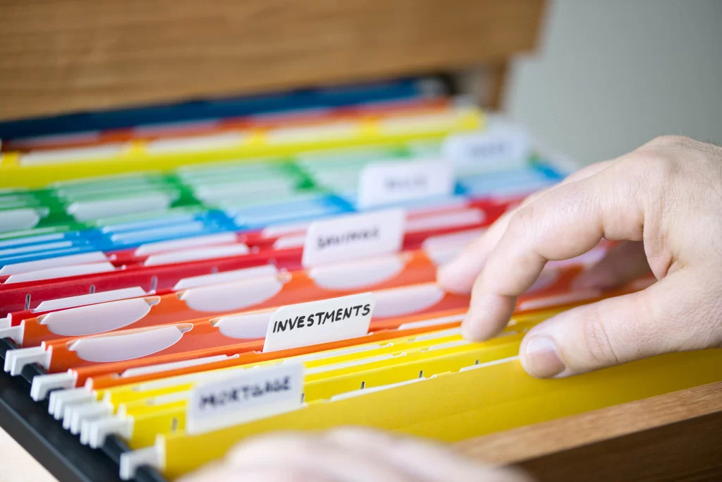 A hand searching through rainbow filing folders. One label reads "Investments" while the other labels are blurred out