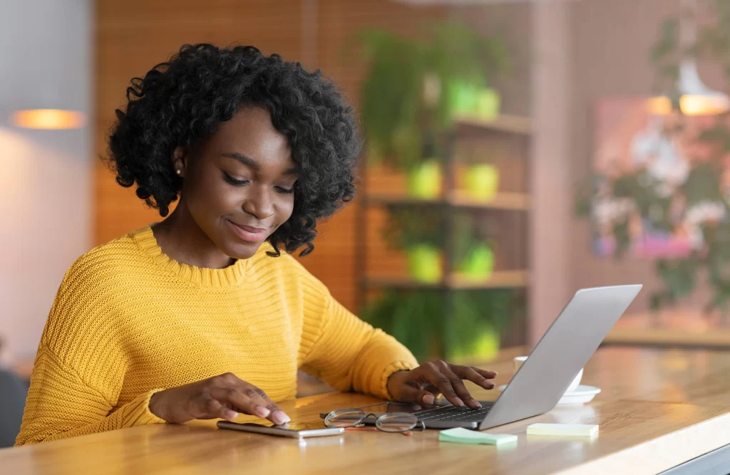 A smiling Black woman working on her phone and laptop