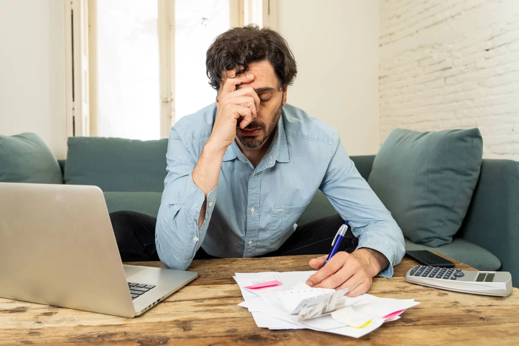 A man with papers and calculator in front of him, holding one hand to his face in frustration