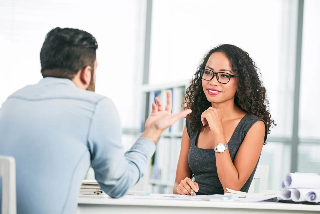 Two professionals talk while sitting at a desk.