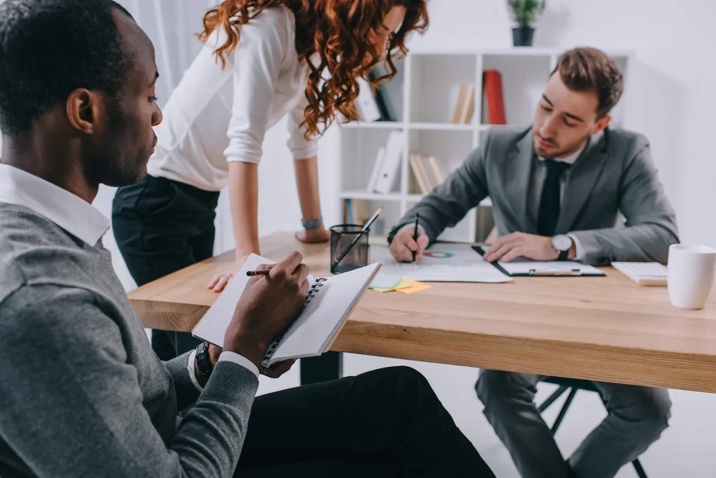 Three professionals work at a table; one is writing in a notebook while another makes notes on a printed graphic.