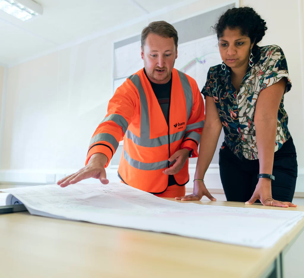 A man in a hi-vis orange jacket talks over blueprints to a woman in an office.