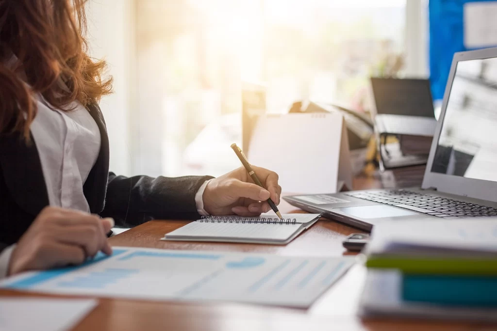 A woman writes in a notebook at a desk covered in papers, files, and a laptop