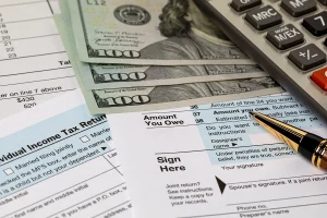 Image includes tax returns, three $100 bills, a pen, and calculator.