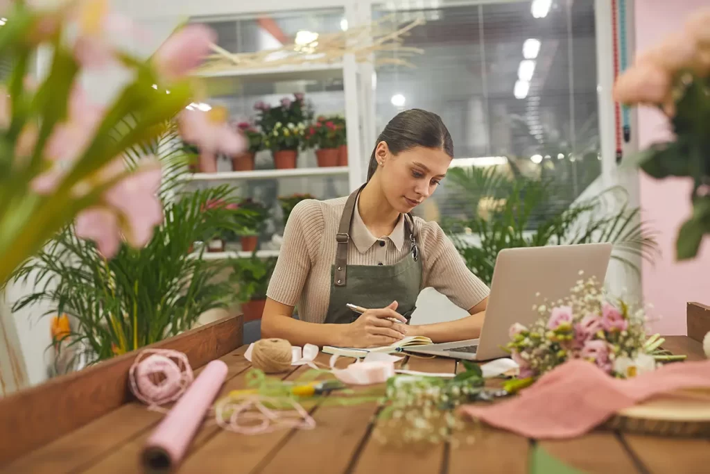 A woman in an apron working on a laptop at a table surrounded by plants