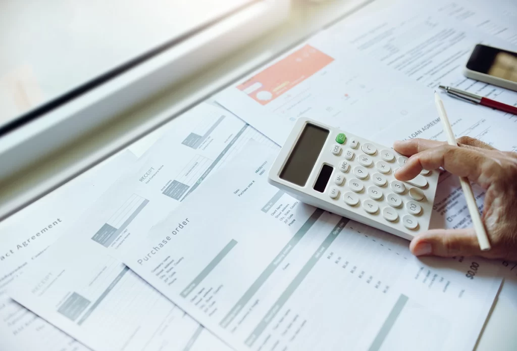 A closeup of a hand typing numbers on a calculator on top of purchase order invoices
