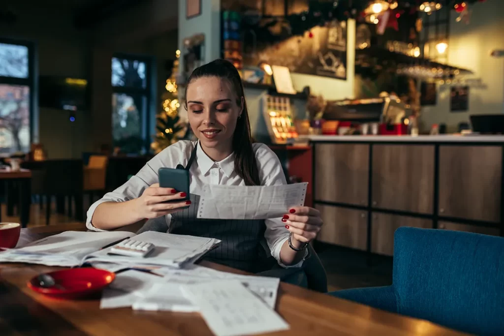 A woman in a closed cafe with a calculator, bookkeeping journal, and papers on the table