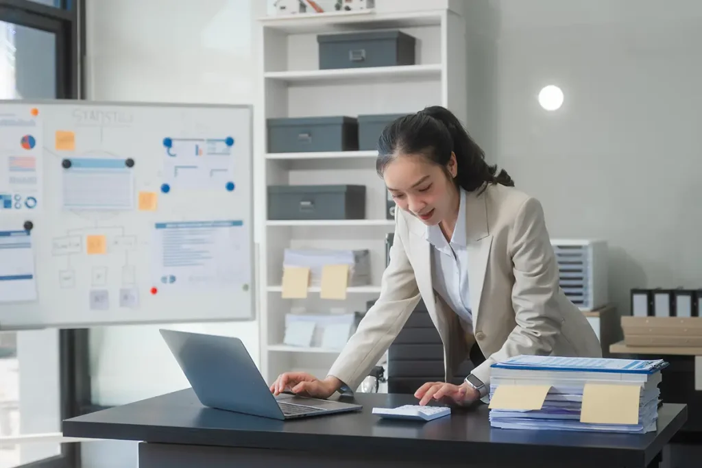 A young Asian businesswoman works alone at a desk with a laptop, stack of papers, and documents
