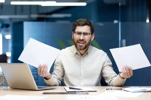 Portrait of angry young male bookkeeper sitting at a desk in the office, holding documents in his hands and shouting at the camera in frustration