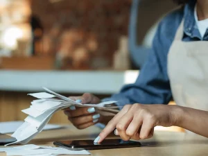 Person reviewing a stack of receipts and using a smartphone calculator at a desk, representing expense tracking or tax preparation.