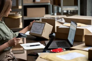 Woman scanning labeled boxes with laptop in front of her.